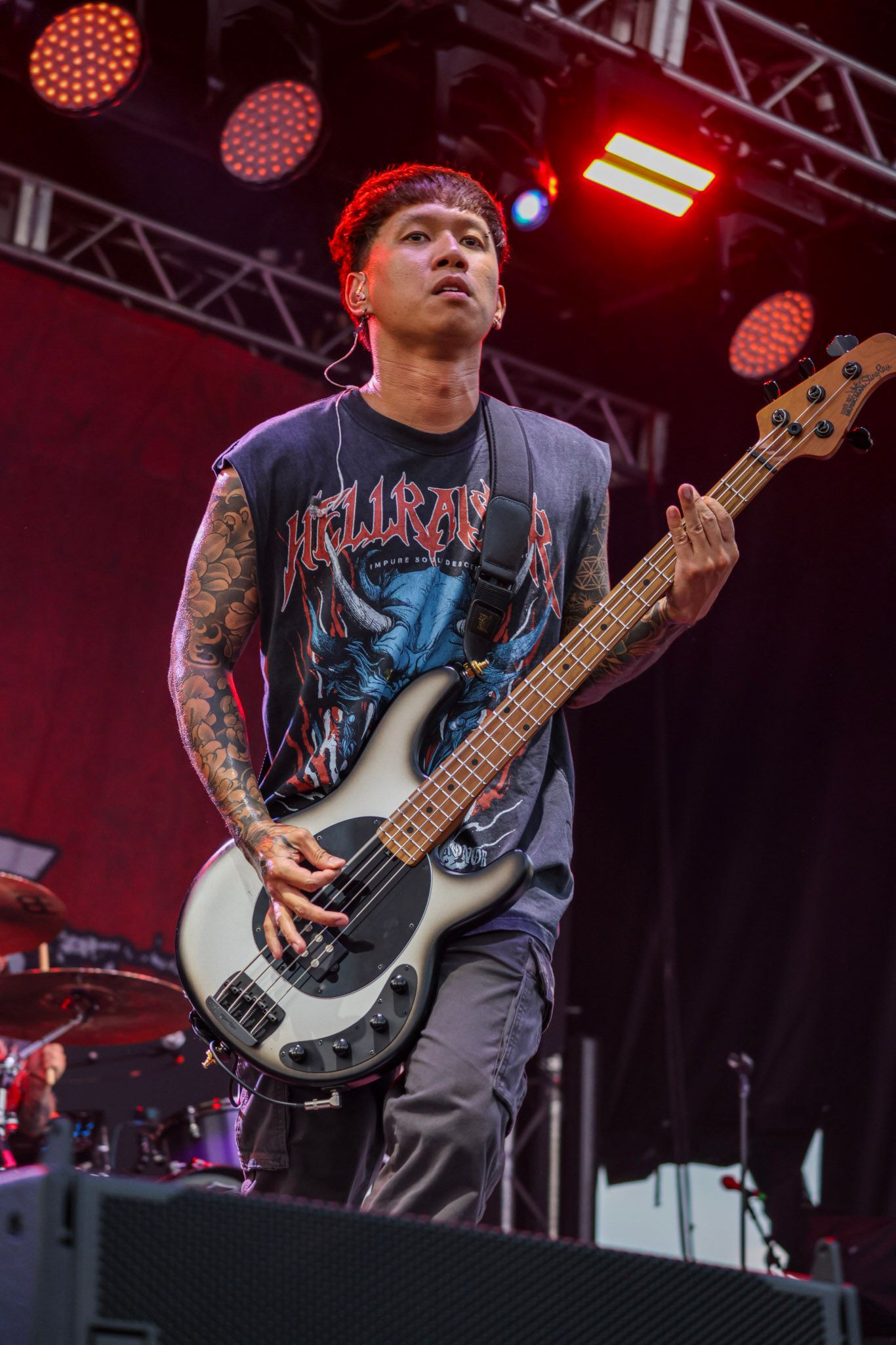 Bass guitarist on stage, tattooed arms, playing a bass guitar. Wearing a band t-shirt. Red and stage lights.