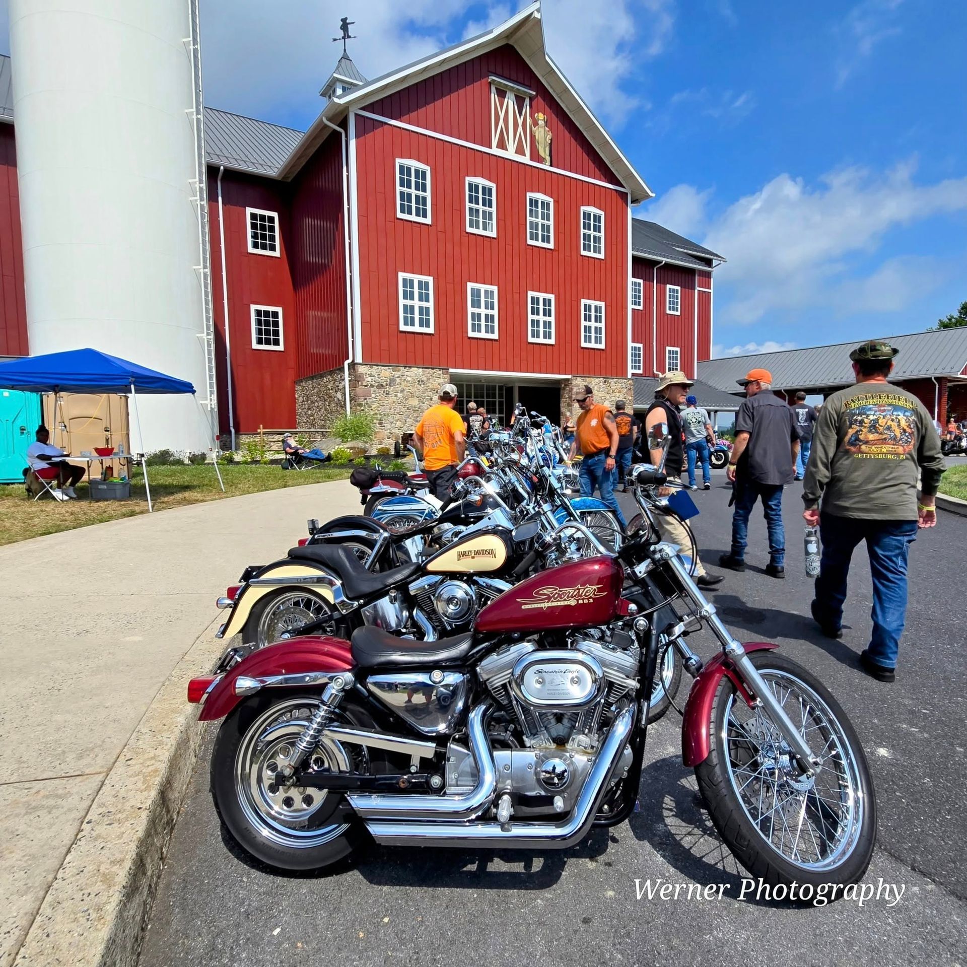 Motorcycles parked in front of a red barn. People gather on a sunny day.