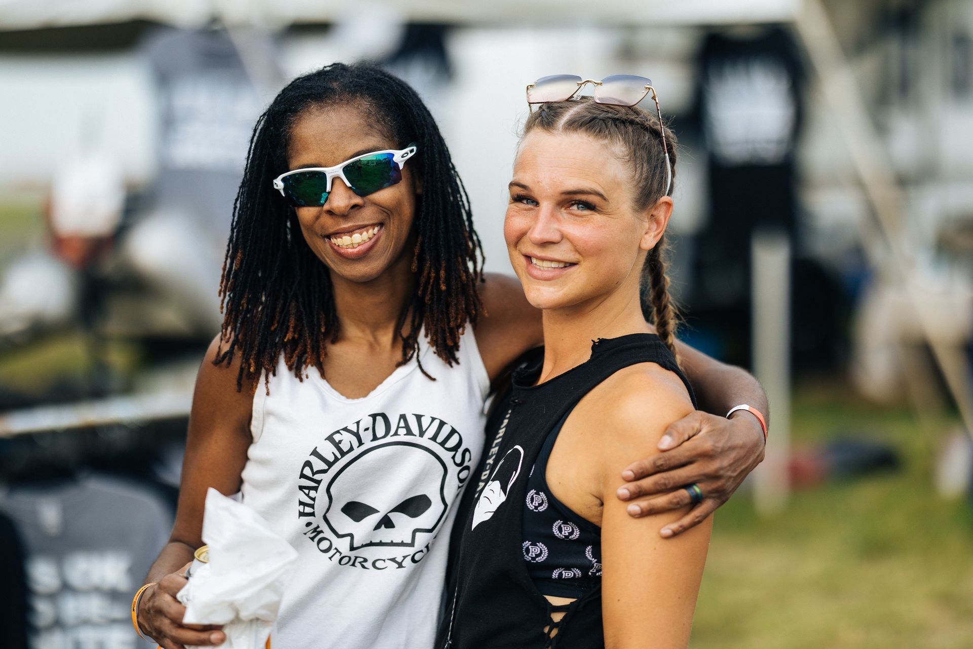 Two women smiling, one with sunglasses and a Harley-Davidson shirt, the other with an arm around her.