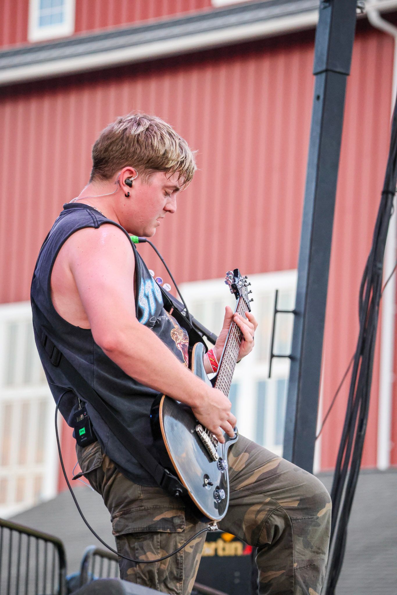 Man plays electric guitar on stage, wearing camouflage pants and sleeveless shirt, red barn in background.