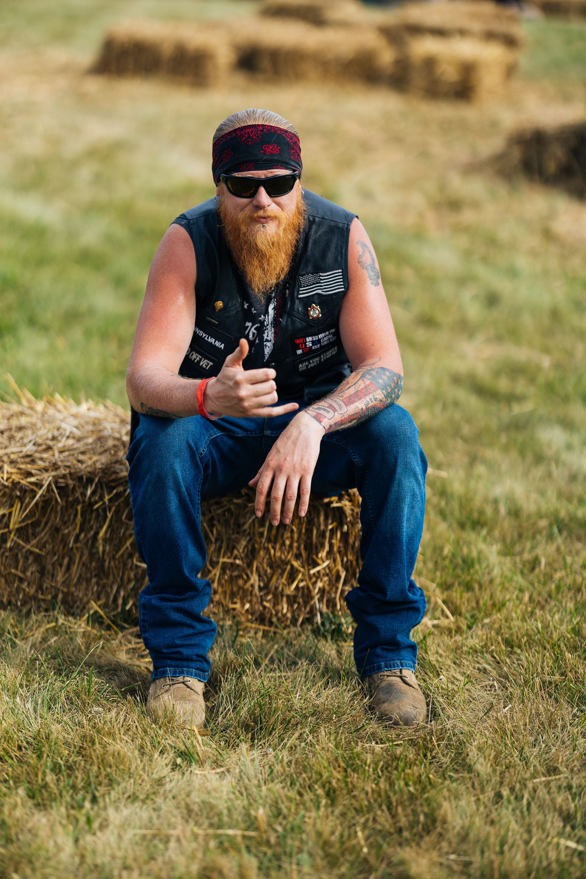 Man with red beard, sunglasses, and bandanna sits on hay bale outdoors, giving a shaka hand gesture.