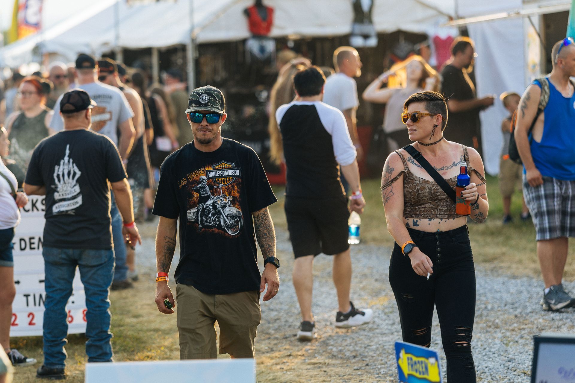 People at an outdoor event, some with tattoos. Man in Harley-Davidson shirt walks. Woman with drink and arm tattoos.