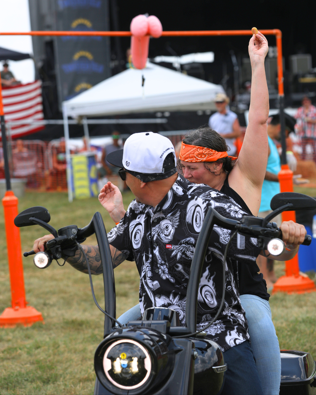 Two people on a motorcycle at an outdoor festival, aiming for a pink object hanging from a structure.