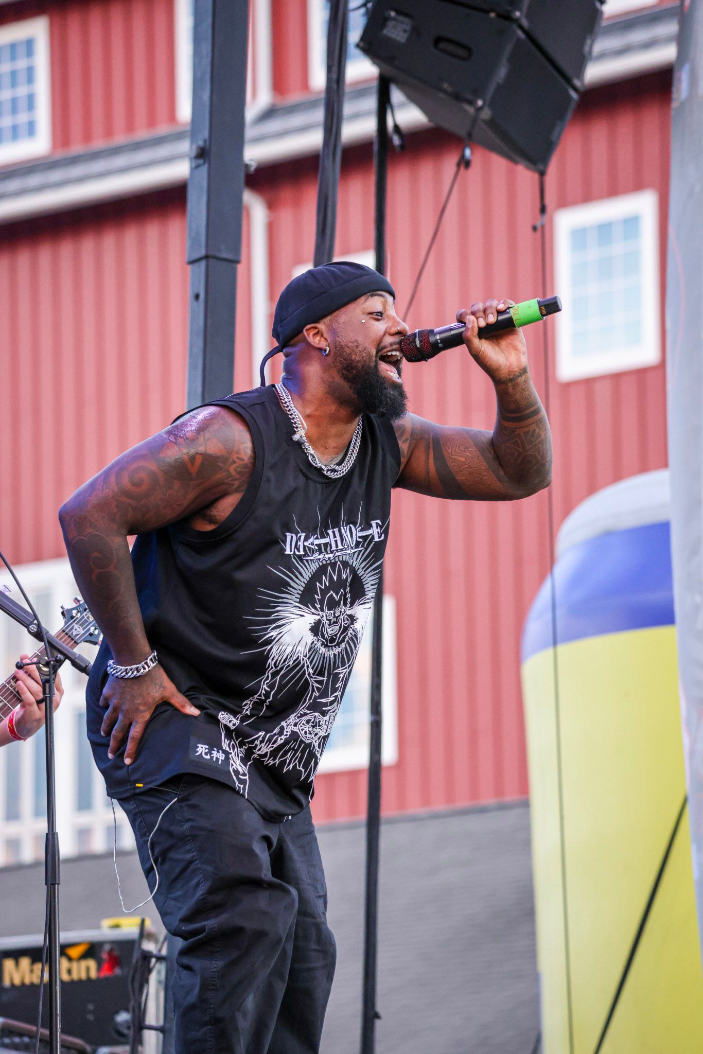 Man with tattoos sings into a microphone on a stage with a red building backdrop.