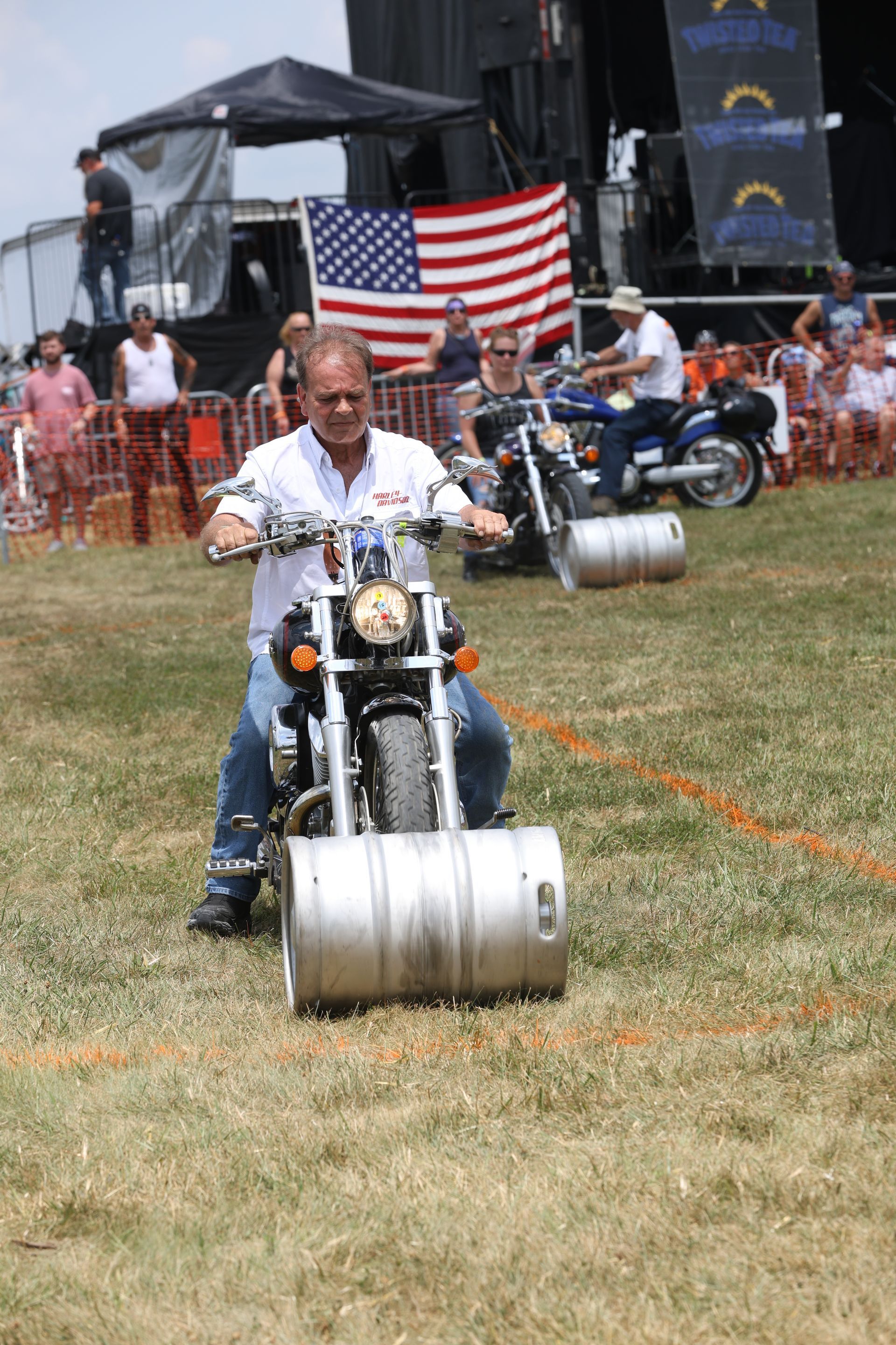 Man on motorcycle with metal barrel front wheel, racing on grass; American flag background.