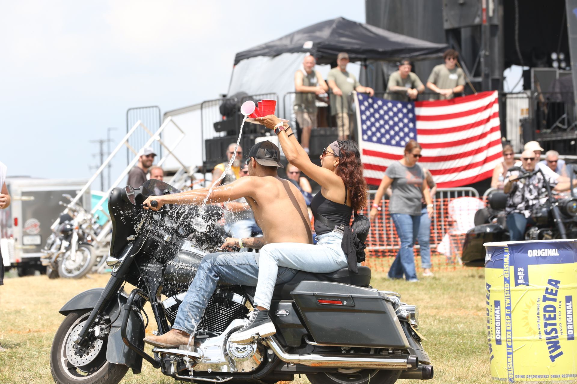 Couple on motorcycle, splashing water, in front of a stage with a flag and crowd at an outdoor event.