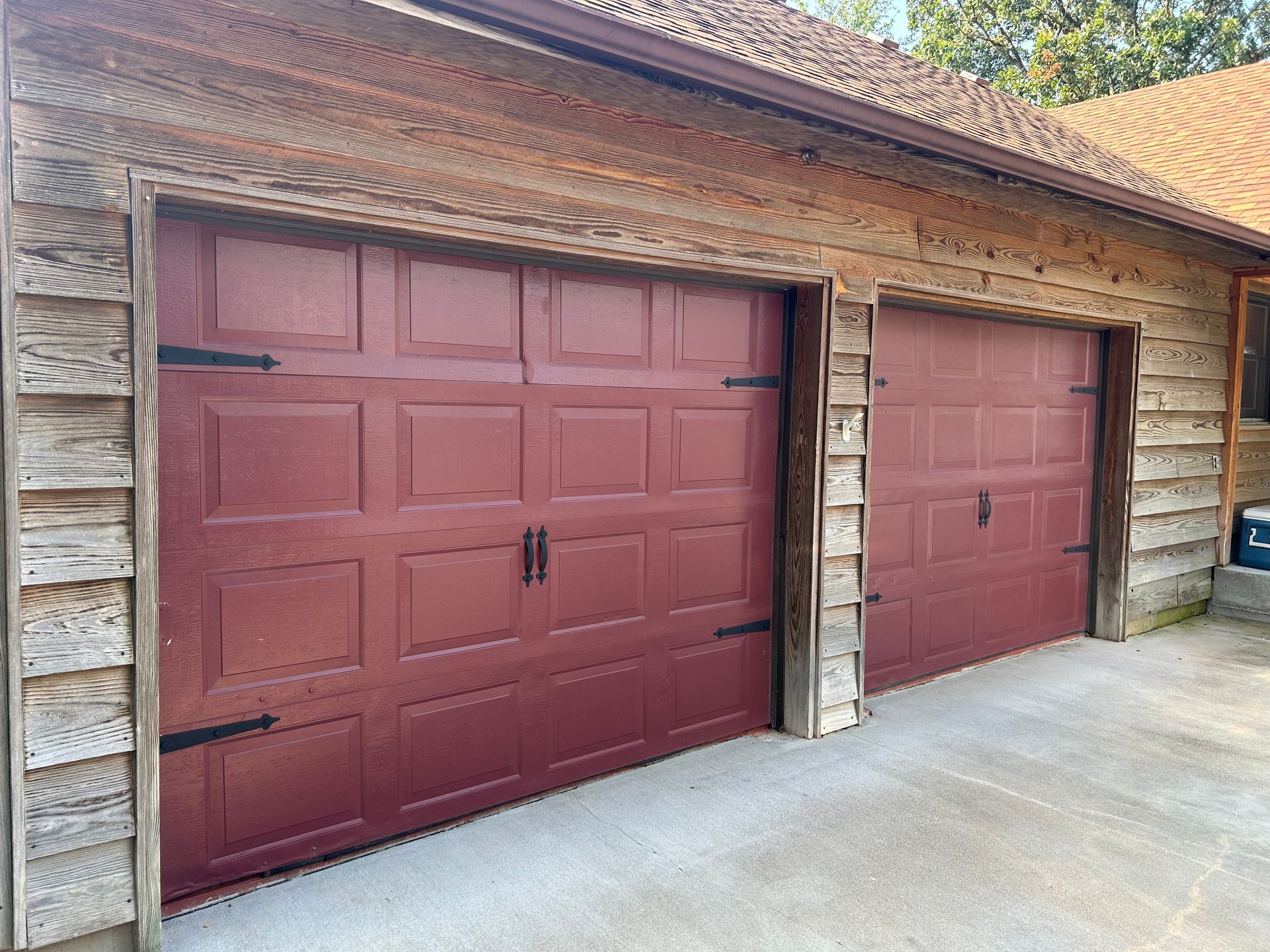 Two maroon garage doors with black hardware, wooden siding, and concrete driveway.