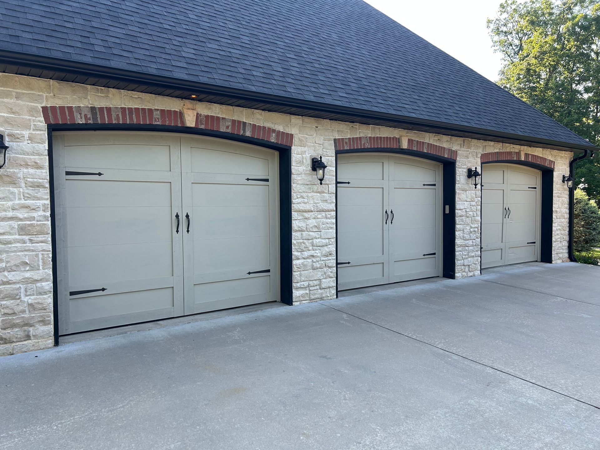 Three-car garage with tan doors, brick trim, and stone facade under a dark roof.