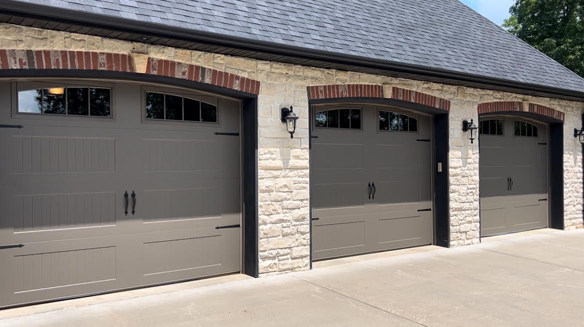 Three-car garage with gray doors, arched brick trim, and stone facade under a dark roof.