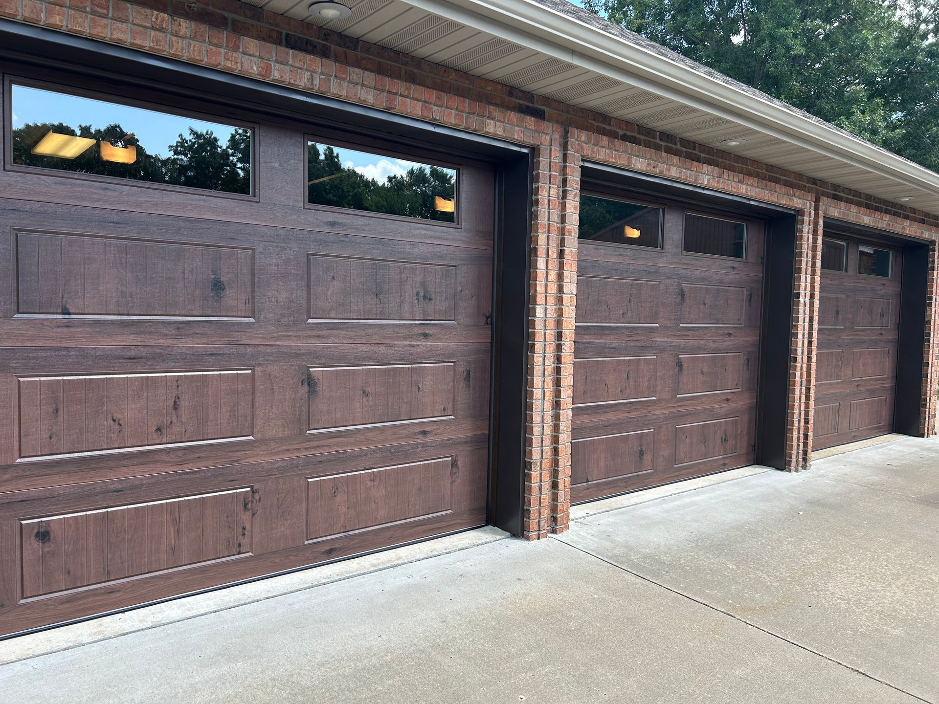 Three brown garage doors with brick accents. Reflective glass windows above the doors. Concrete driveway.