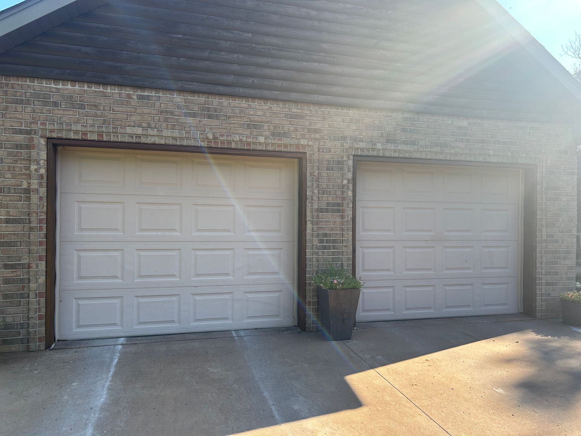 Two beige garage doors in a brick building; a potted plant sits nearby.