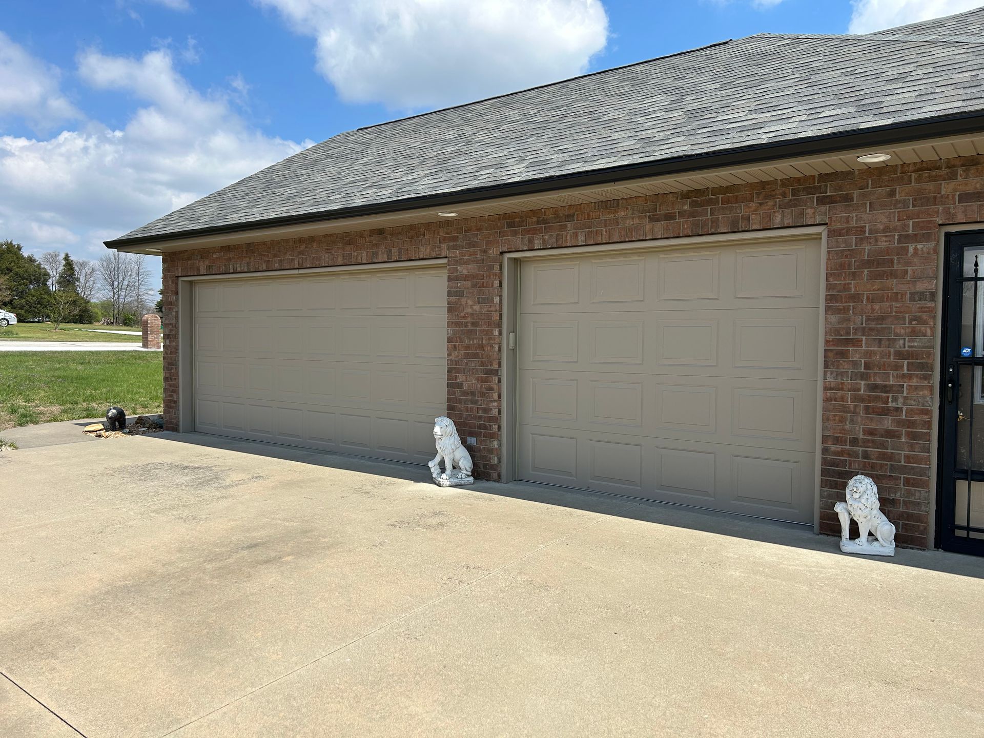 Two-car garage with beige doors, red brick exterior, and concrete driveway. White lion statues flank garage doors.