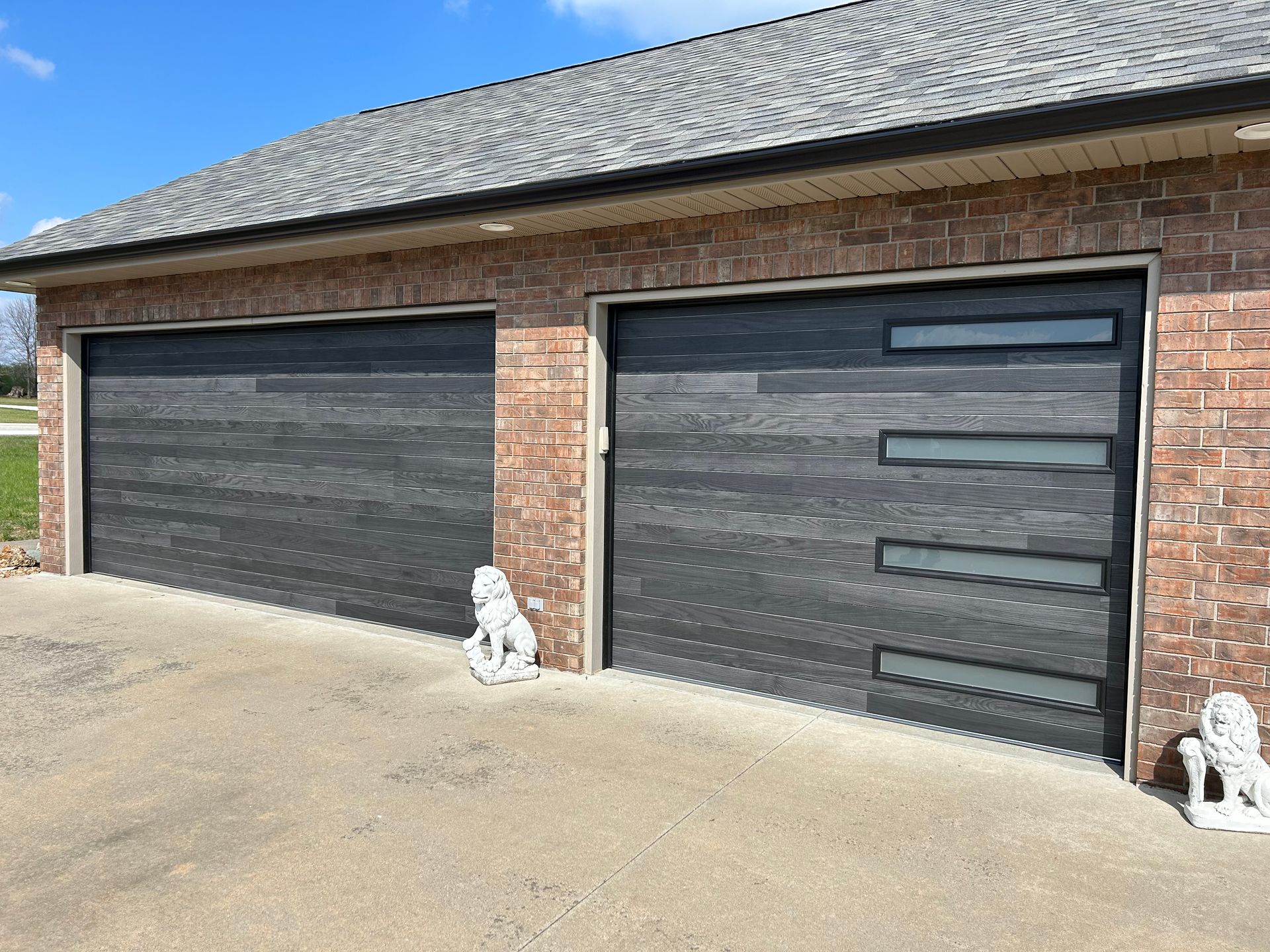 Two dark gray garage doors on a brick building with decorative windows and concrete driveway.