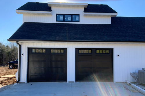 Three olive-green garage doors with windows, in front of a tan house. Concrete driveway with brick border.