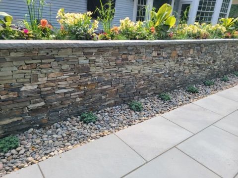 a stone wall surrounded by gravel and flowers in front of a house