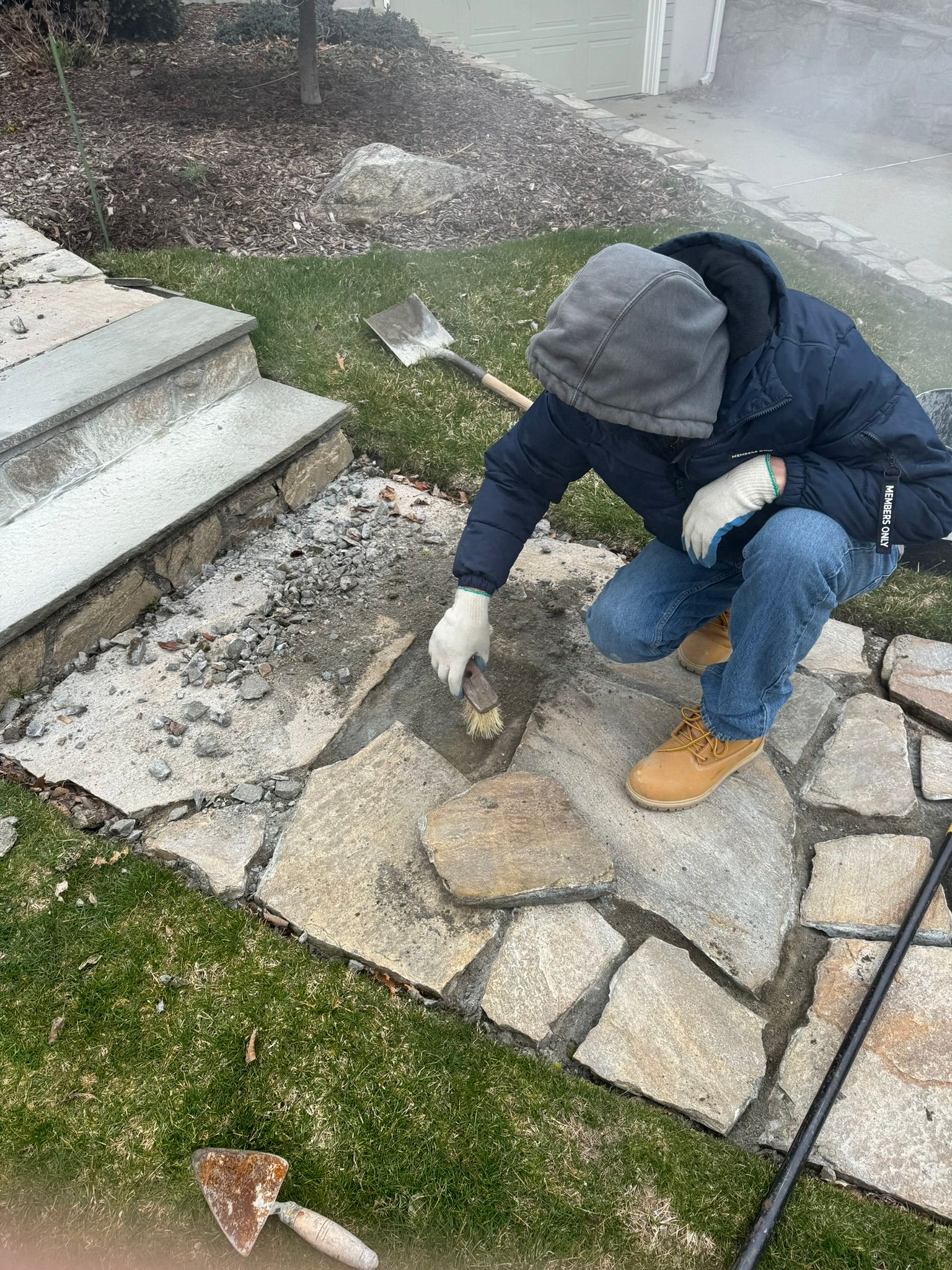 A man is kneeling down on a stone walkway with a shovel