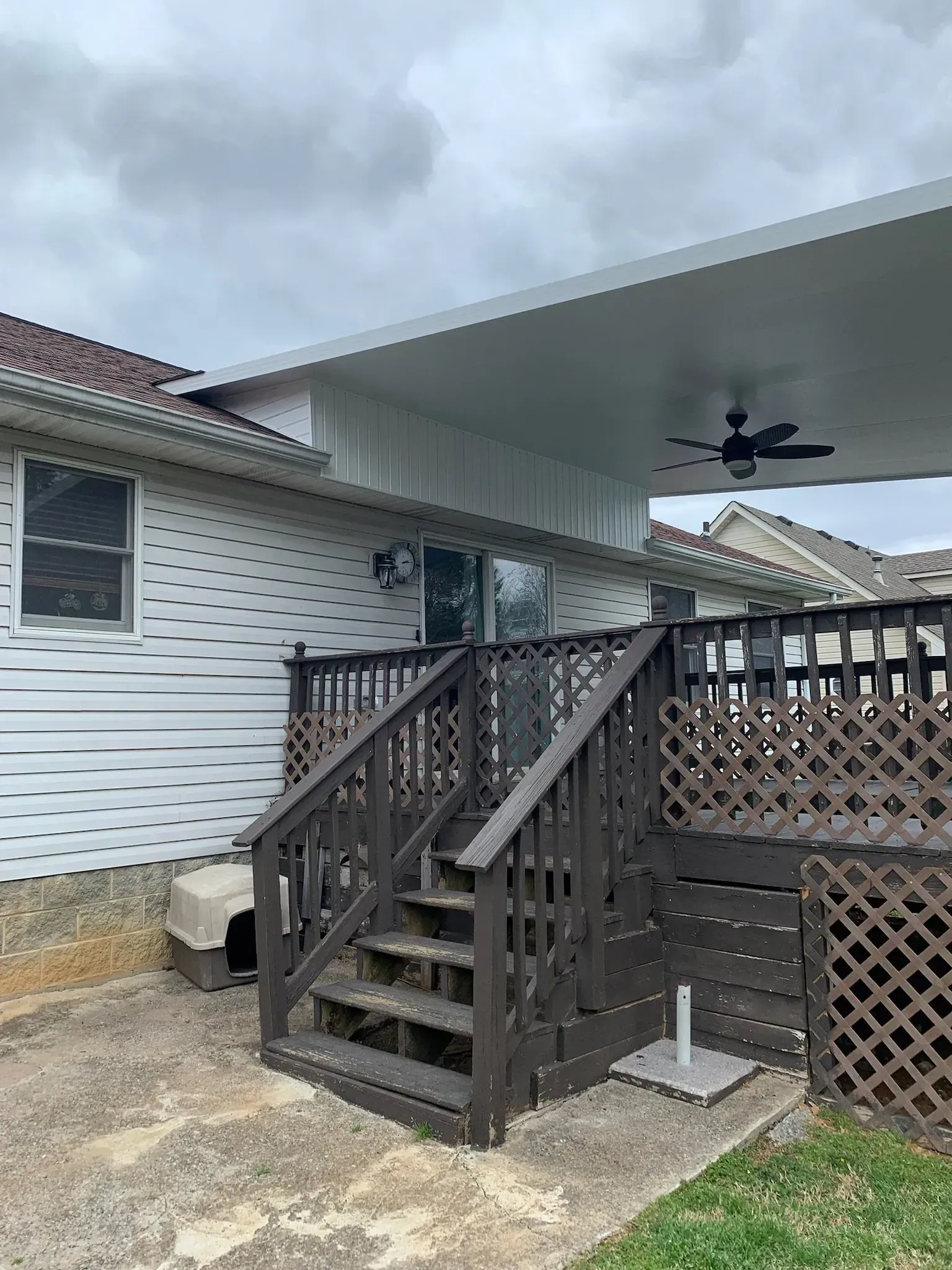 Brown deck with stairs, leading to a covered patio attached to a white-sided house, under a cloudy sky.