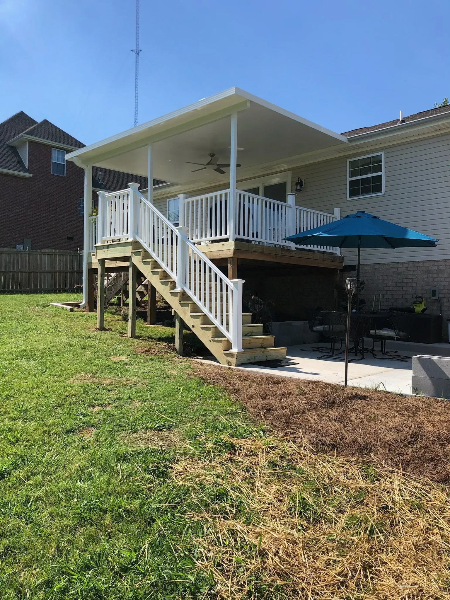 A raised deck with a covered patio, white railing, and stairs, next to a house with a blue umbrella.