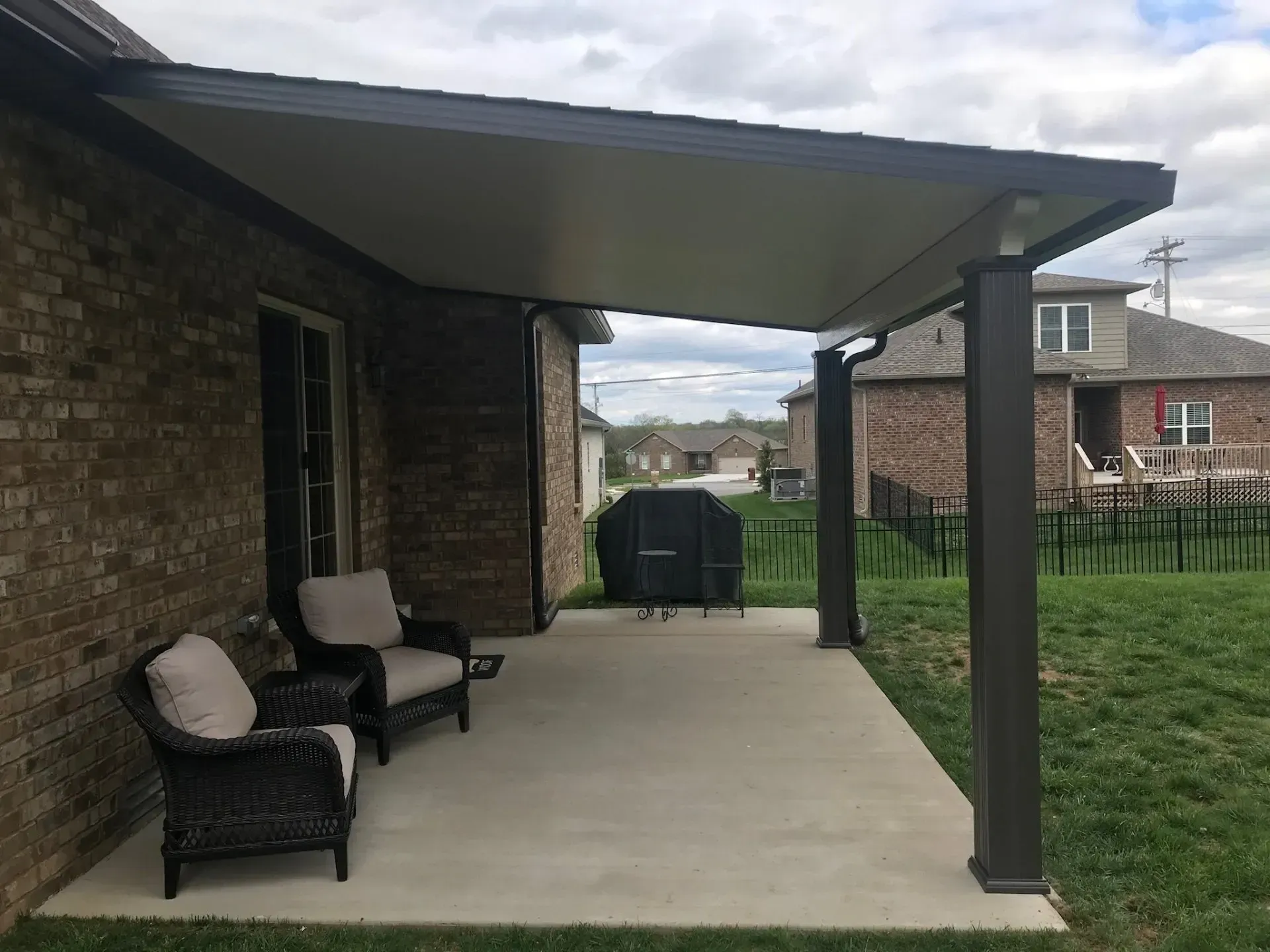 Patio with dark gray awning, chairs, and grill next to a brick house.