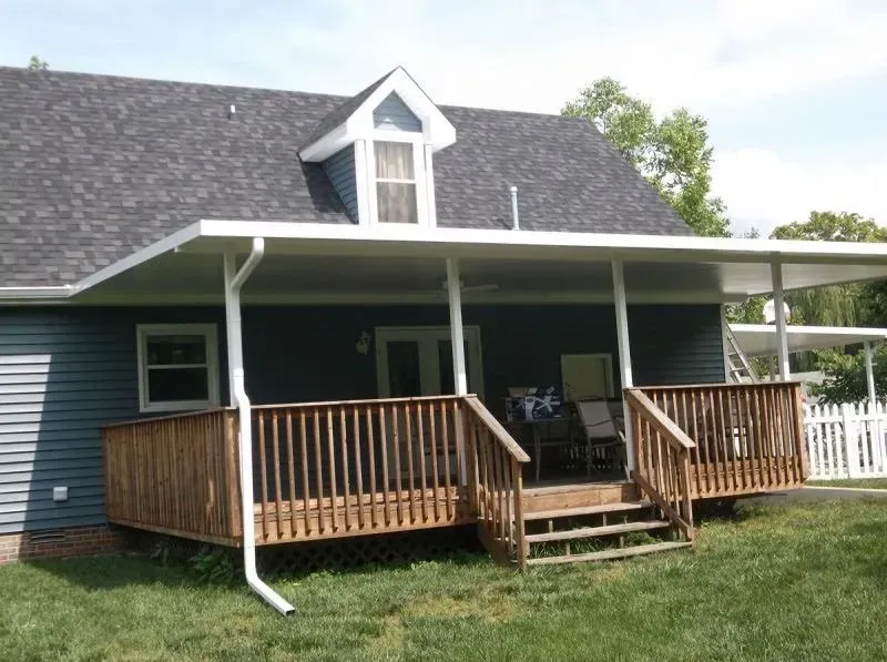 Wooden deck with white-railed awning attached to a blue-sided house with a dormer and gray roof.