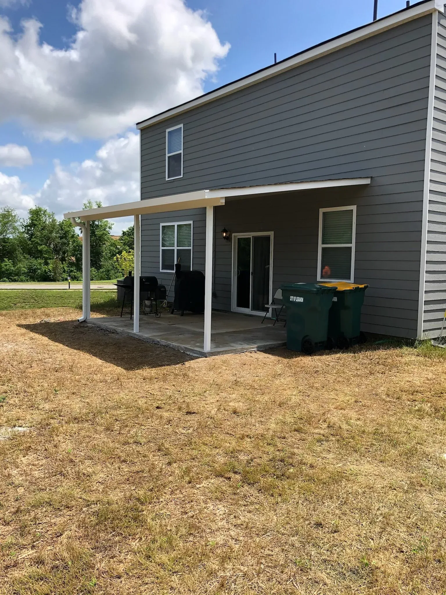 Back of a gray house with a white patio cover. Two green trash bins are visible next to a sliding glass door.