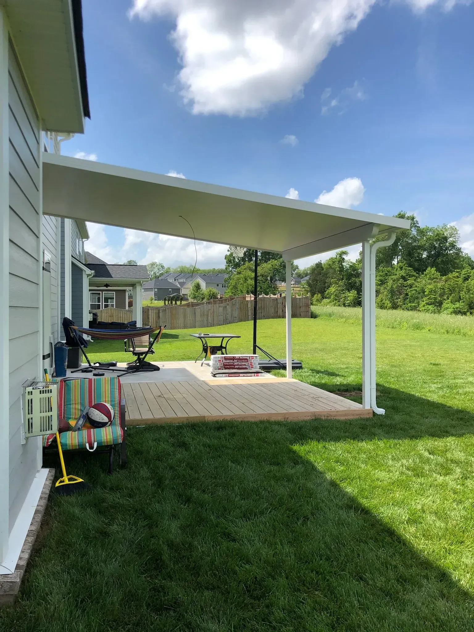 A white patio cover extends from a light blue house over a wooden deck, overlooking a grassy yard.