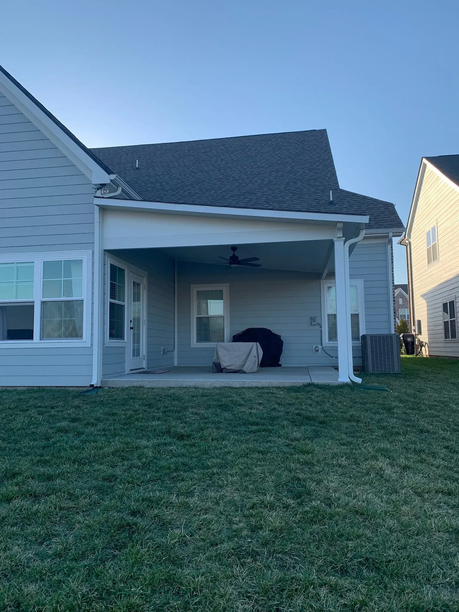 Backyard with light blue house, covered patio, green grass, and clear sky.