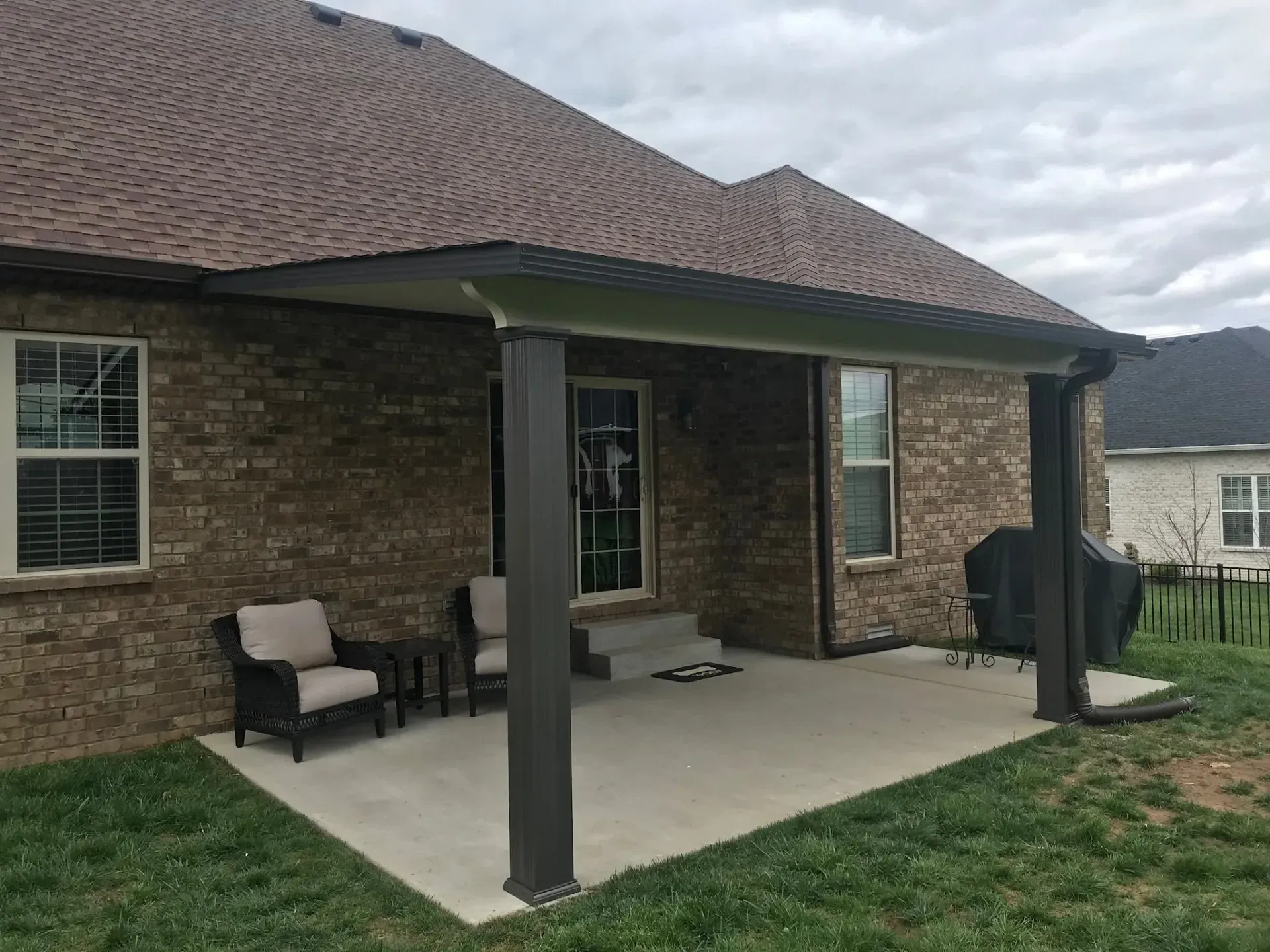 Back patio with concrete floor, brick house, and brown roof. Two chairs and a table are on the patio.