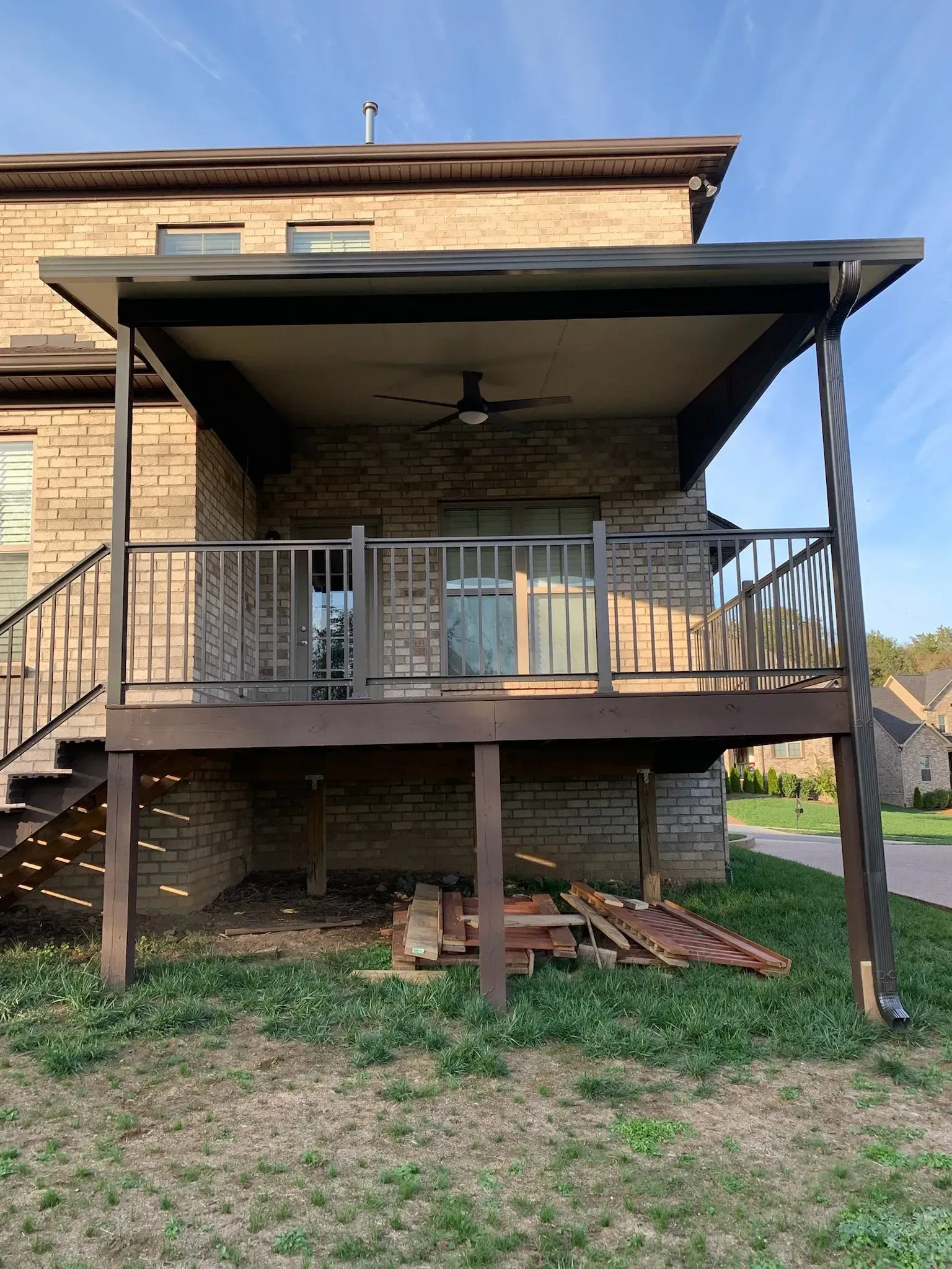 Brown deck with a covered patio, attached to a brick house, overlooking a grassy yard under a blue sky.