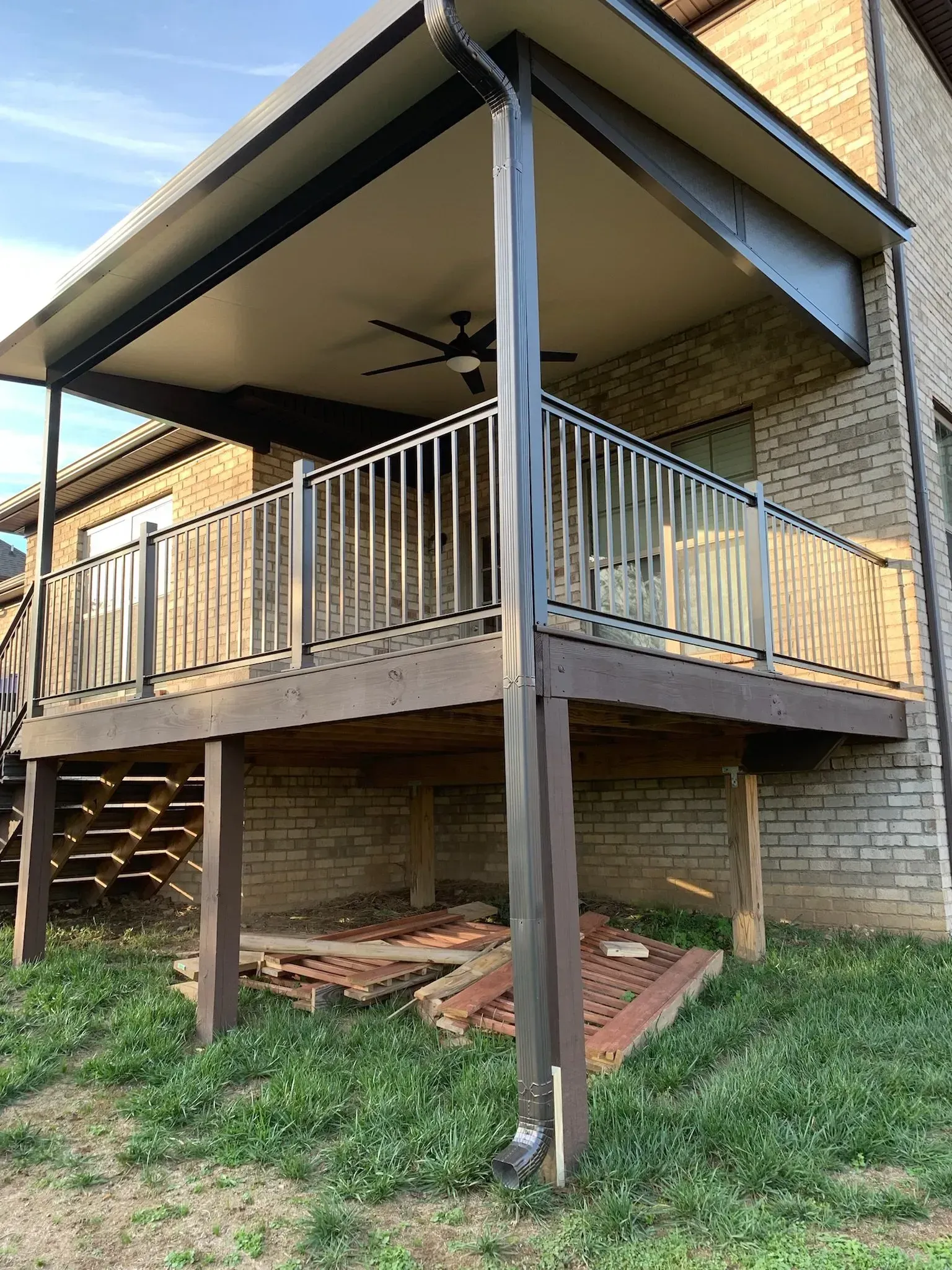 Deck with brown railing, metal posts, and covered ceiling, attached to a brick house.