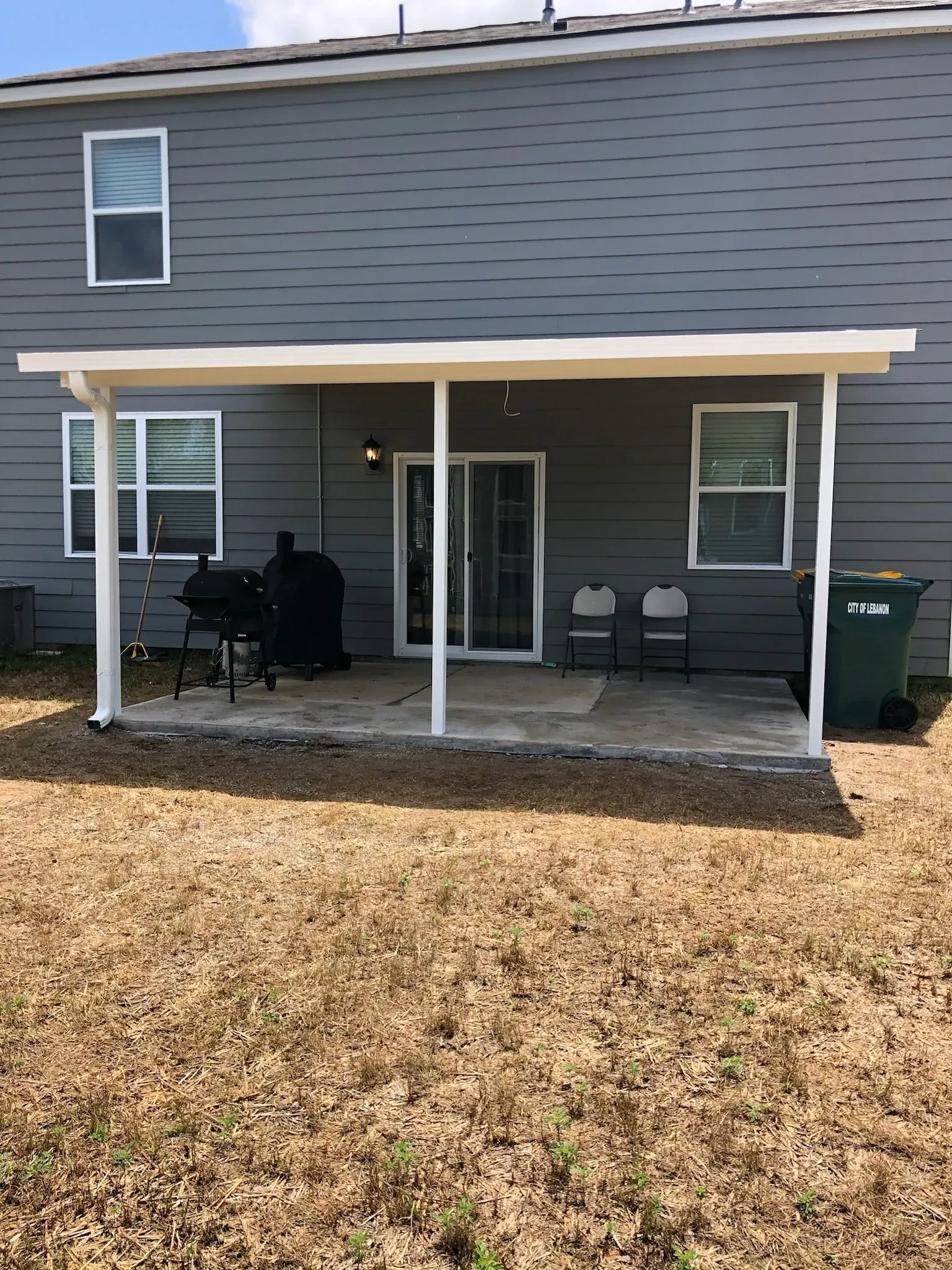 A patio with an attached covered awning. A grill sits next to a sliding glass door and two chairs.
