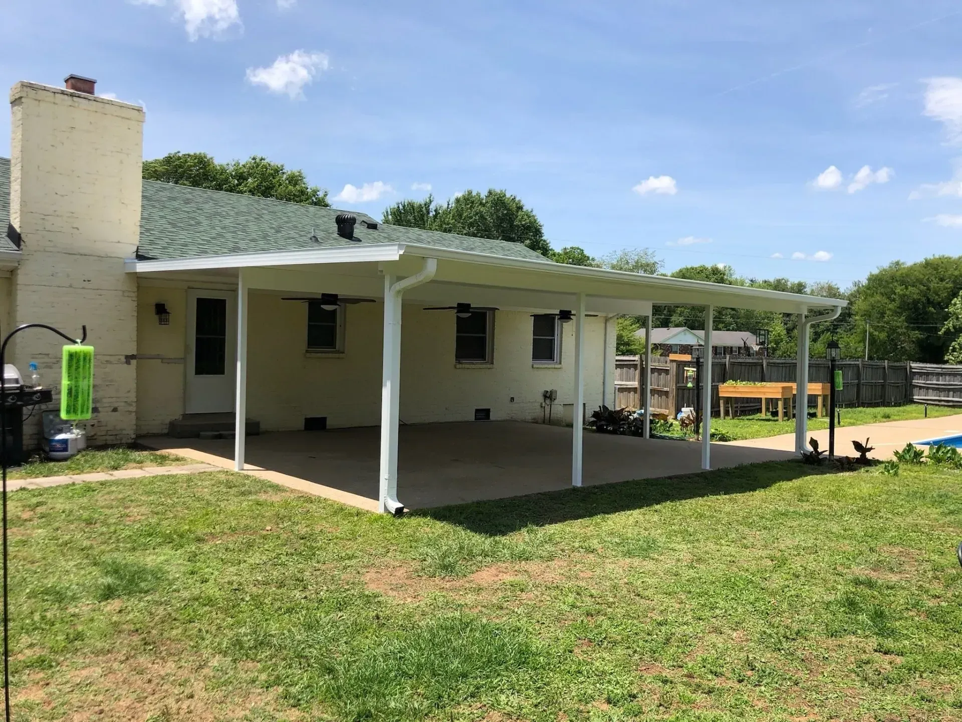 A white-columned patio cover extends from a light-colored house over a concrete patio. Green grass surrounds the patio.