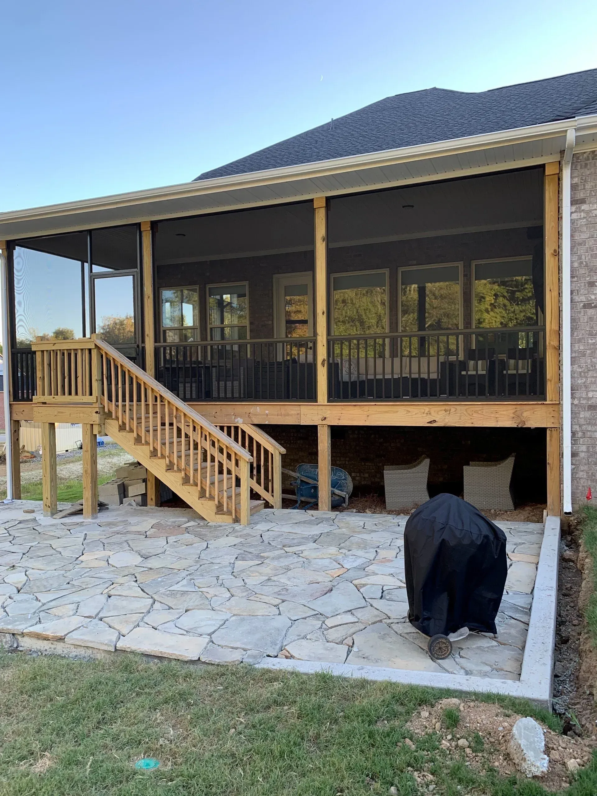 Screened-in porch with wooden stairs, overlooking a stone patio. A grill is covered, and the sky is blue.