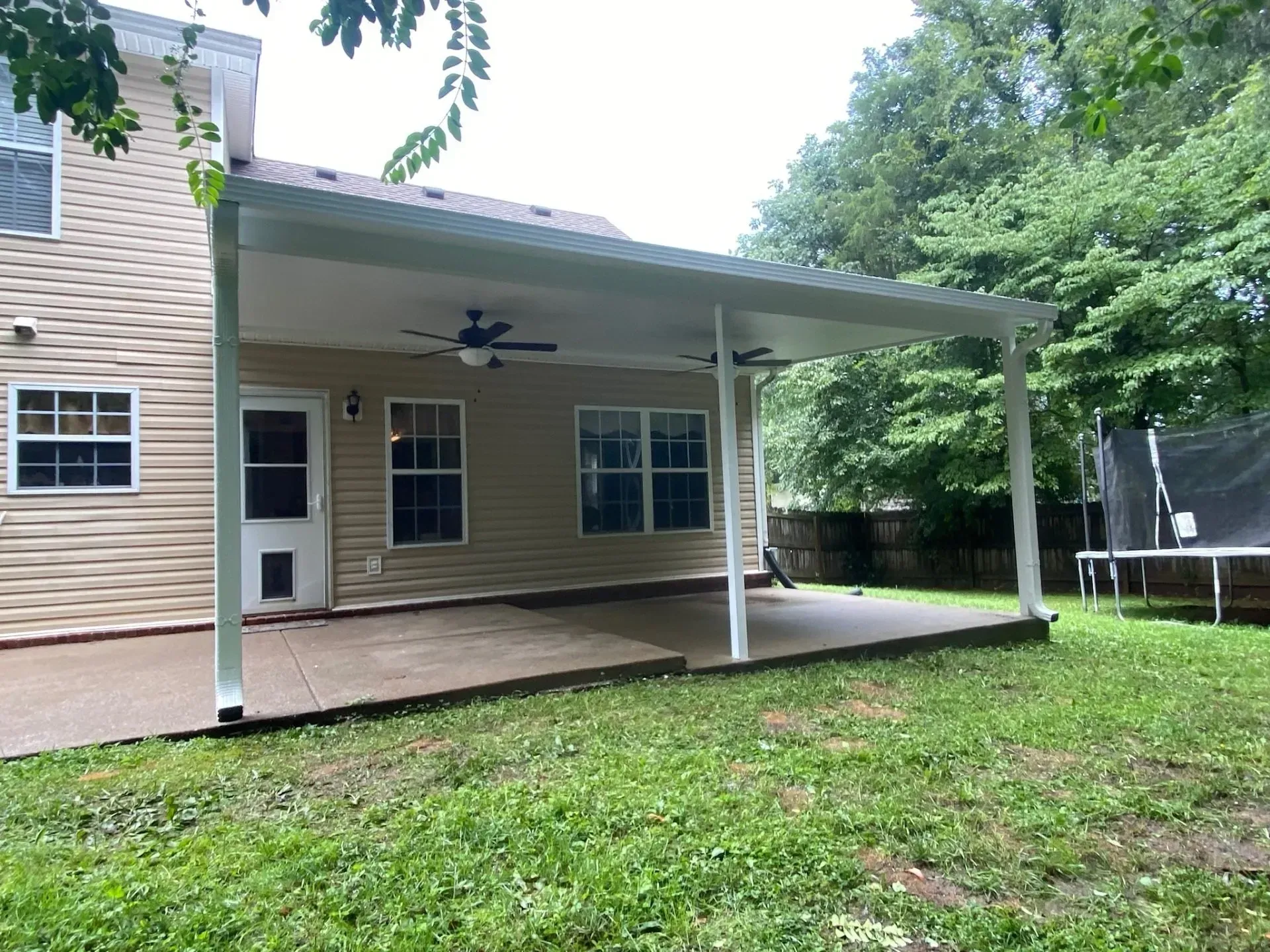 Covered patio attached to a house with a concrete floor, fan, and columns. Green yard.