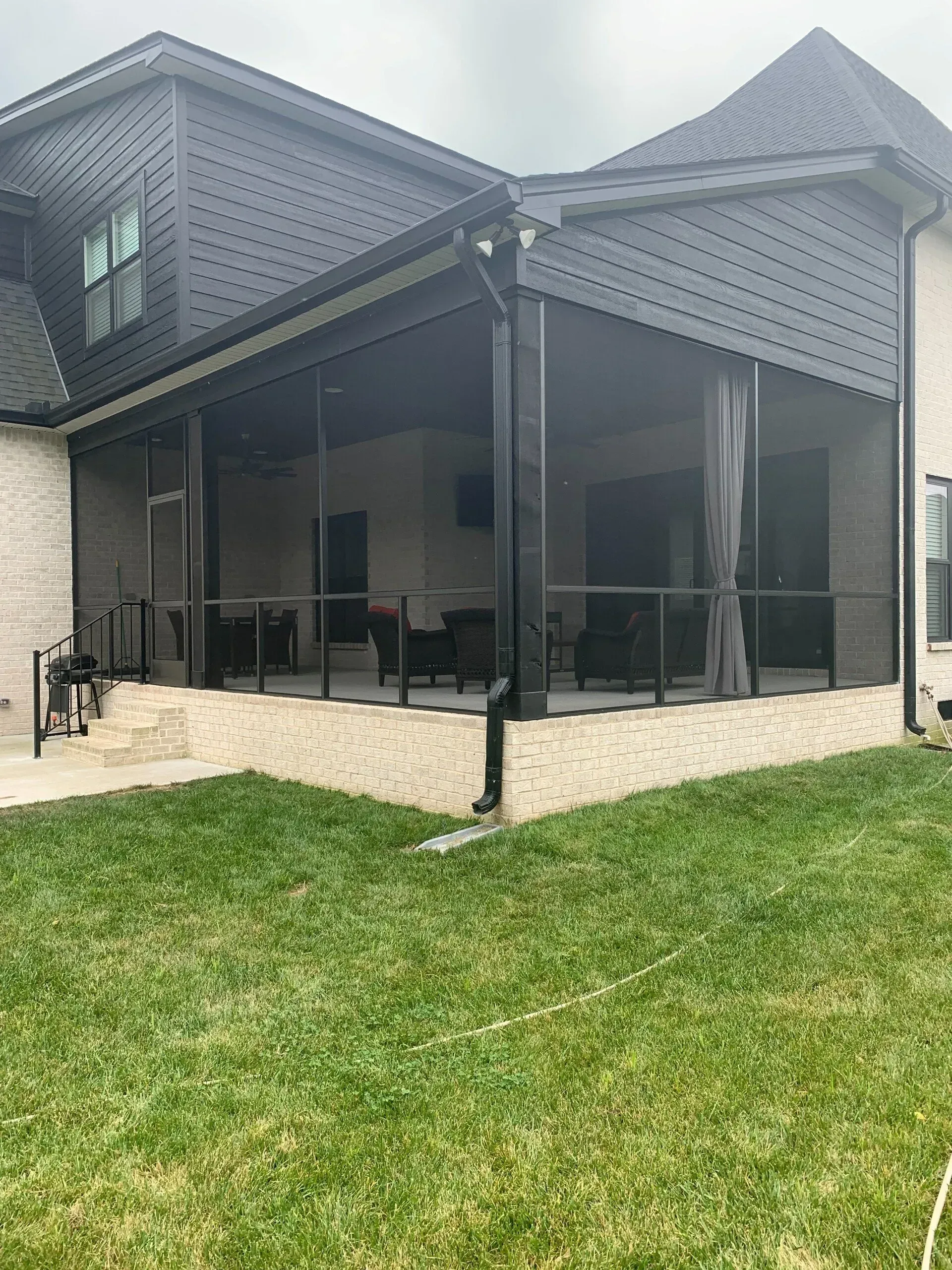 Screened-in porch on a brick house. Black screens, railings, and roof. Grass in foreground. Overcast sky.