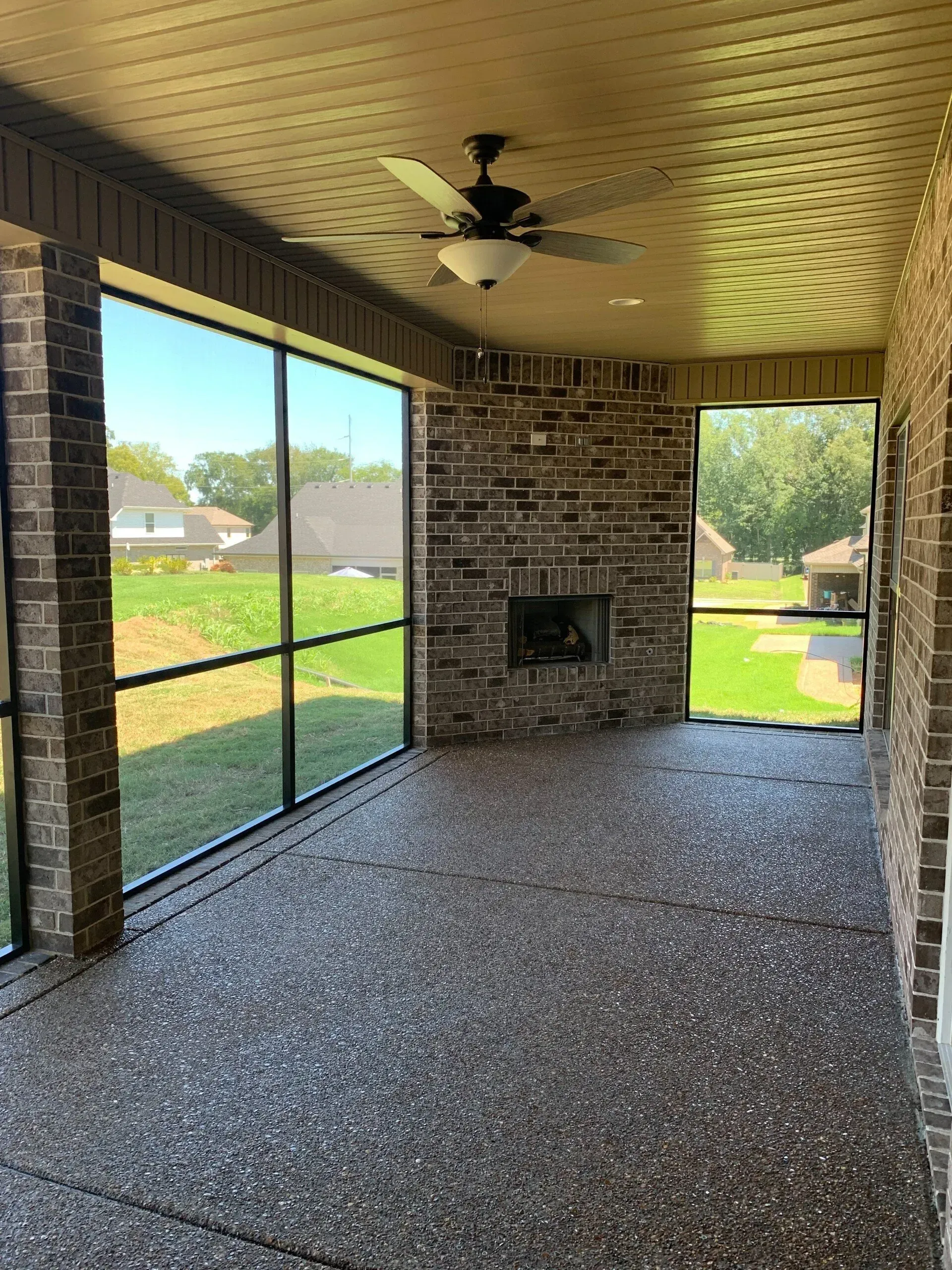 Screened-in porch with brick fireplace and textured flooring, windows overlooking a green yard, and a ceiling fan.