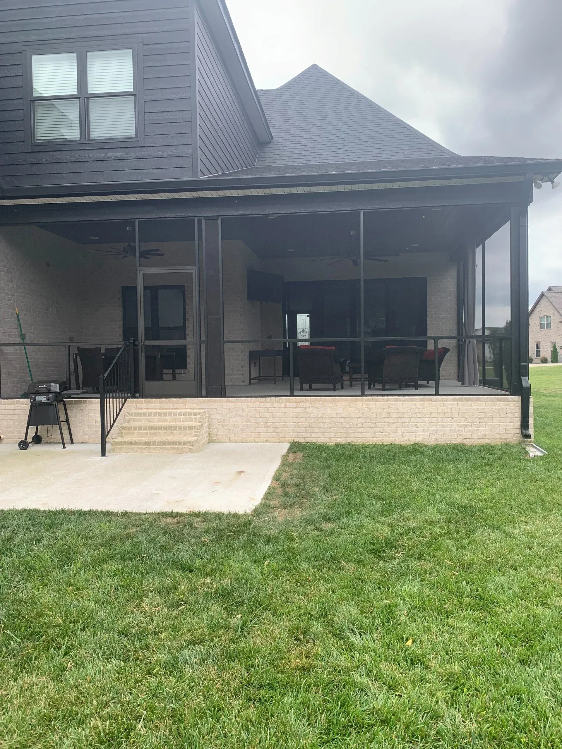 Screened porch with dark trim, outdoor furniture, and a grill, adjacent to a house with a shingled roof.