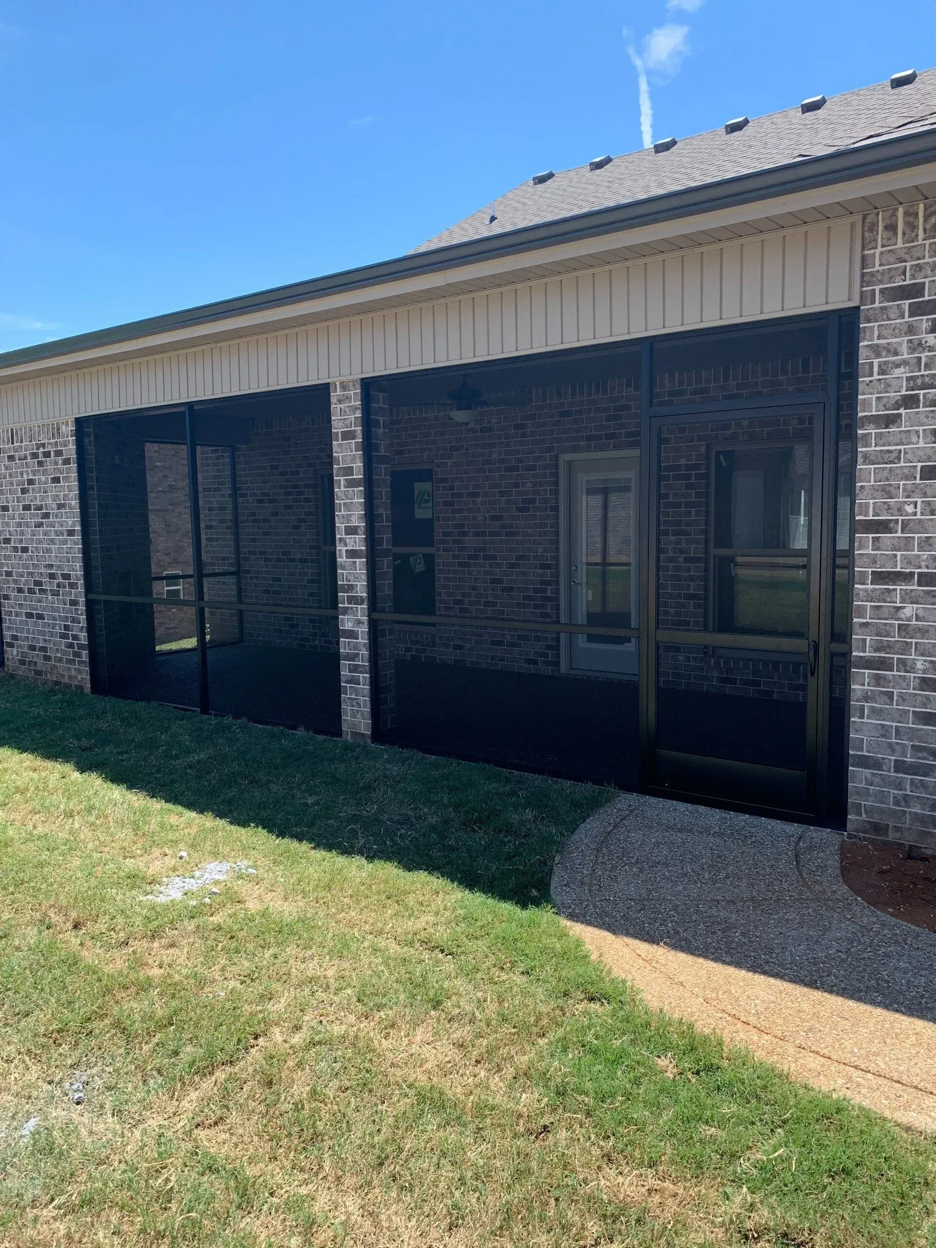 Screened-in porch on a brick house. Black screens, tan siding, green grass, and gravel path. Sunny day.