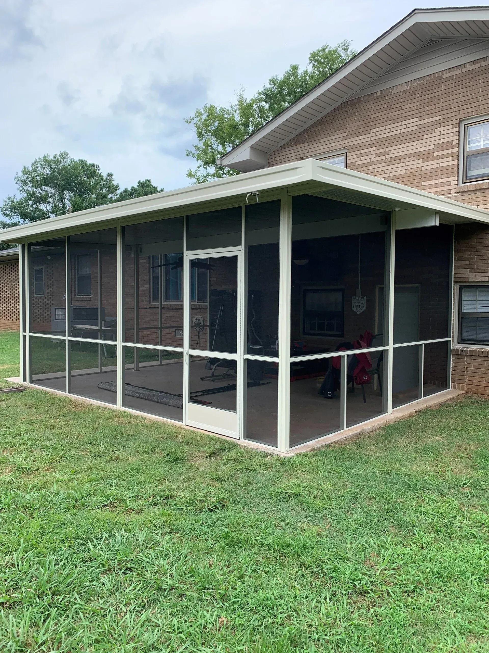 Screened-in porch attached to a brick house with white trim. Green grass surrounds it. Cloudy sky in the background.