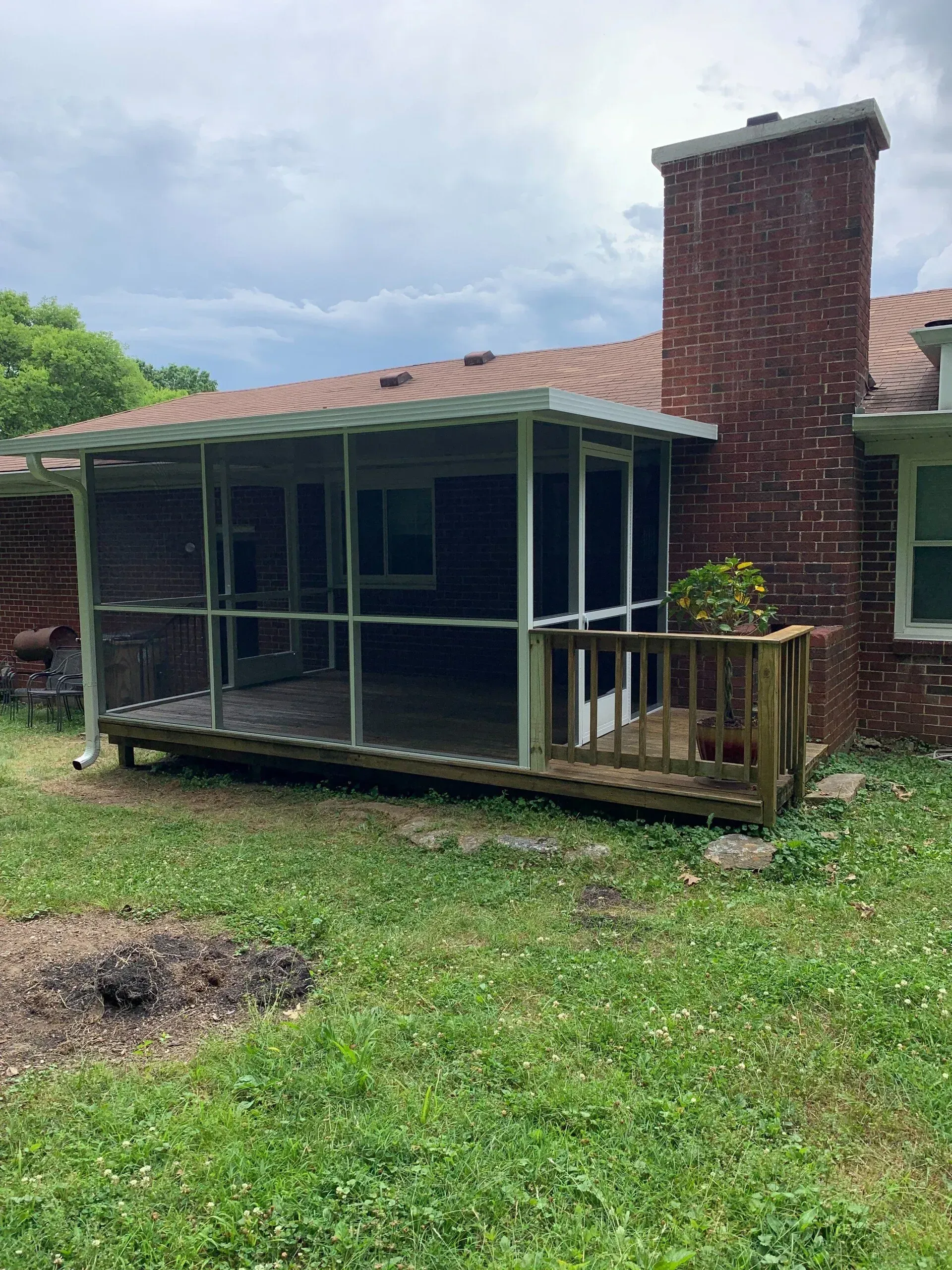 Screened porch attached to a brick house with a chimney and a small wooden deck. Overcast sky.