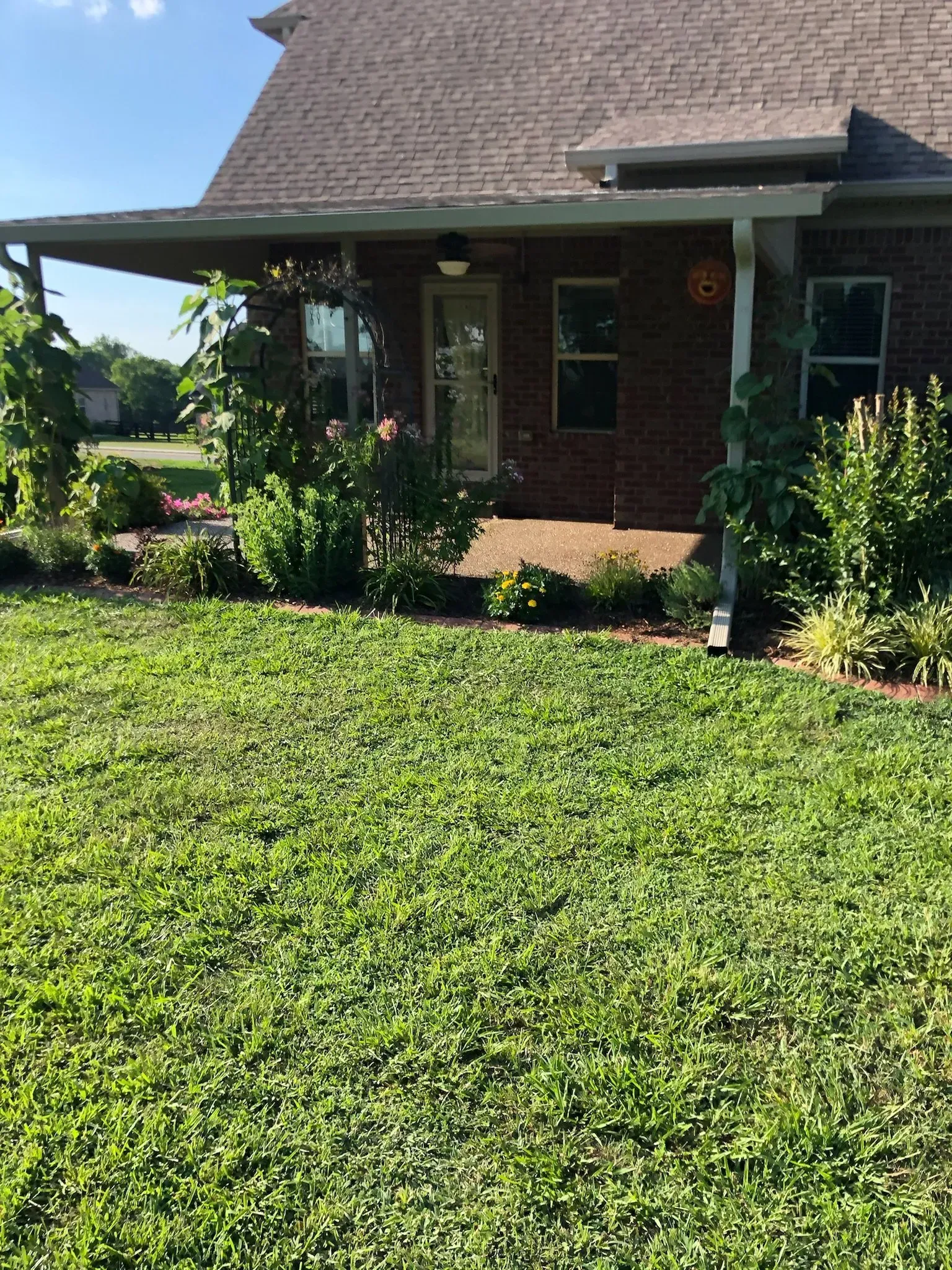 A brick house with a covered patio, garden beds, and green lawn.