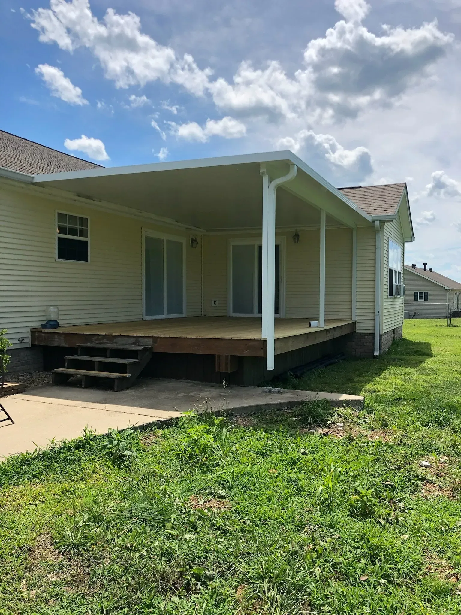 Covered patio attached to a beige house, with deck, sliding doors, and green grass.