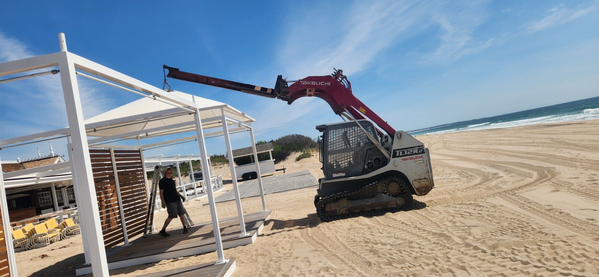 A skid steer loader on a beach is lifting a white gazebo, person nearby, blue sky and ocean in background.
