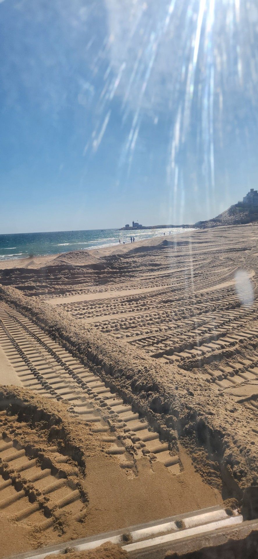 Beach scene with blue sky, sand, ocean. Tire tracks in sand. Bright sunlight.