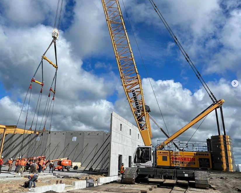 Construction crane lifting a concrete wall panel. Workers on the ground. Bright blue sky with clouds.