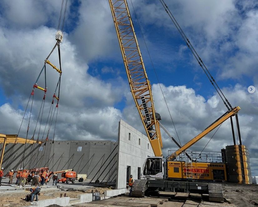 Construction crane lifting a concrete wall panel. Workers on the ground. Bright blue sky with clouds.