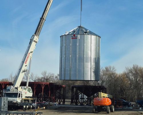 Crane lifting a metal grain silo. Construction site with other structures, blue sky.