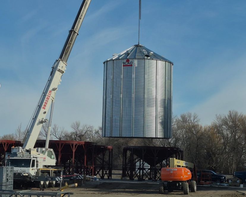 Crane lifting a metal grain silo. Construction site with other structures, blue sky.