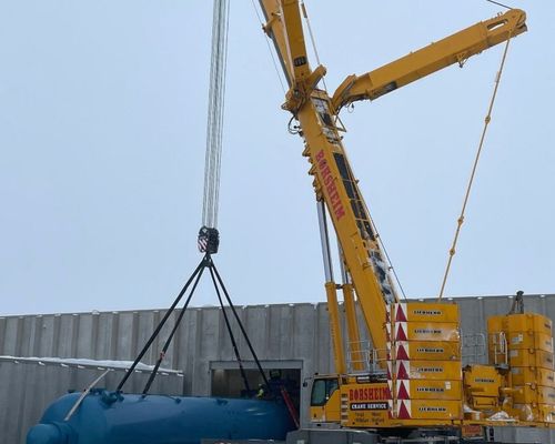 A large crane lifts a blue tank over a building's opening against a cloudy sky.