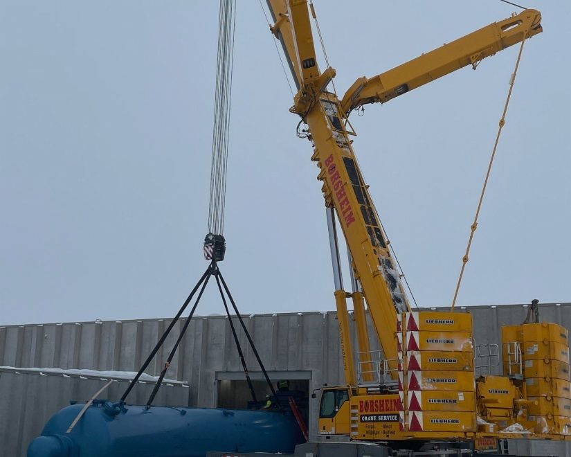 A large crane lifts a blue tank over a building's opening against a cloudy sky.